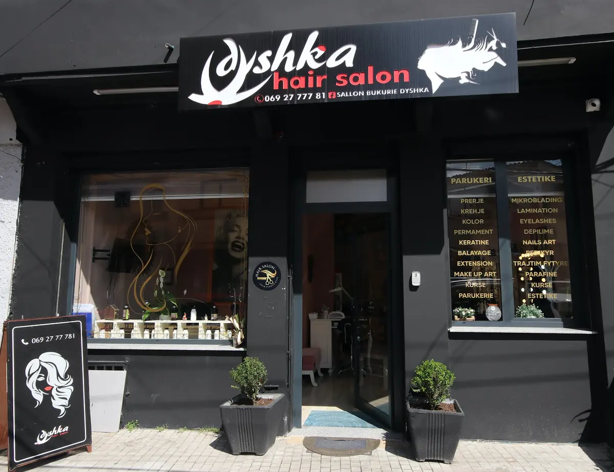 The black shopfront of Dyshka hair salon: white script sign, open door, a window lettered in gold with the service list, and a pavement board with the phone number